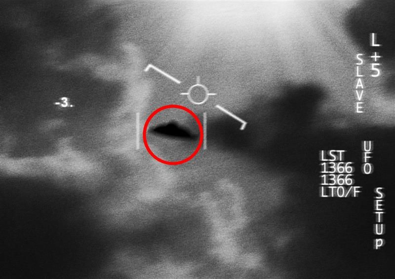 A blurred black and white photo that looks like a military camera tracking UFO in the sky. The saucer-shaped object is in the middle of the image surrounded by clouds. There is a red circle around it and markers on the frame to indicate its position.  