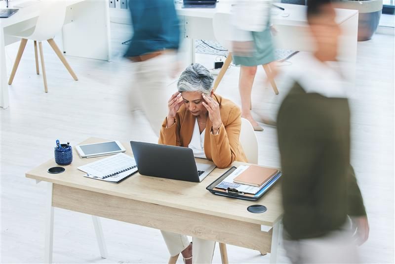 A middle aged woman sits at her desk with her hands pressed to the sides of her head. She is wearing a mustard colored jacket and a white shirt. Her desk has an open laptop in front of her and clipboards and paperwork to either side. There are other people walking around in the image, their bodies are blurred to give the impression that the world is moving quickly while the woman is struggling to focus. 