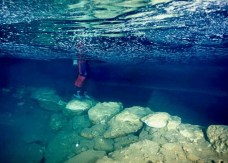 underwater bridge, photographed from just below the surface; someone's legs are visible as they stand on the stones of the bridge