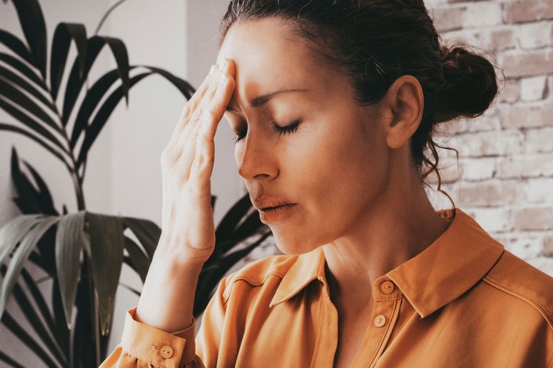 woman with eyes closed holding hand to her forehead to indicate fatigue; she is wearing an orange blouse and there's a brick wall and a plant in the background