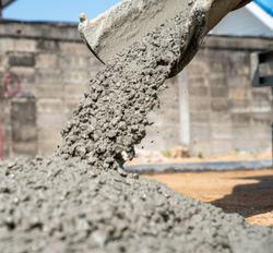 A close up photo of concrete being poured from a mixer. The concrete is piled up as it flows from the tip of a funnel. In the background is a large grey wall. 