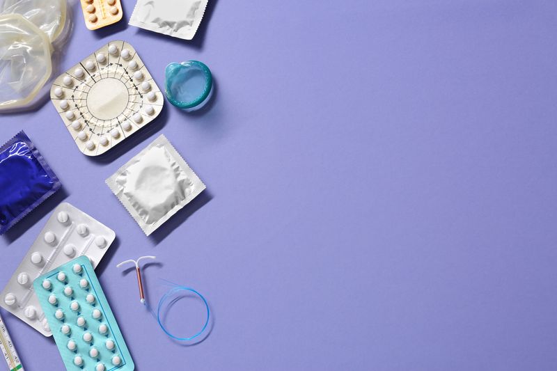 purple background with various contraceptives arranged towards the left hand side: pills, condoms, and an intrauterine device.