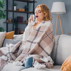 woman sneezing on sofa wrapped in a blanket and surrounded by tissues