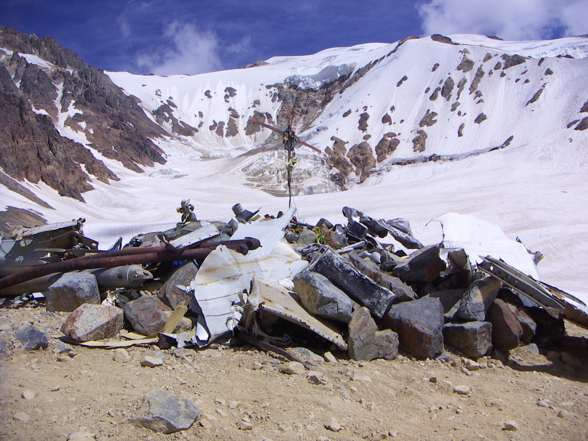 The photo shows the memorial site in the Andes mountain. The memorial consists of rocks and twisted metal left over from the plane that has been piled together. At its centre is a thin cross made of assembled metal. Behind the memorial, the top of the mountain is visible, covered in snow. 