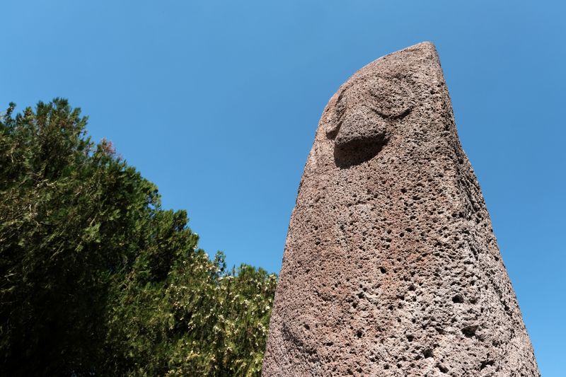 Example of a dragon stone in Armenia. The top of the standing stone is visible with a carved depiction of Vishap, the ancient god of water. There are green tree tops to the left and bright blue sky behind.