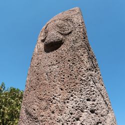 Example of a dragon stone in Armenia. The top of the standing stone is visible with a carved depiction of Vishap, the ancient god of water. There are green tree tops to the left and bright blue sky behind.