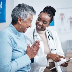 A Black female doctor is explaining information from a clipboard to an elderly lady. They are both laughing and the older lady has her hands pressed together in a similar gesture to a prayer. 