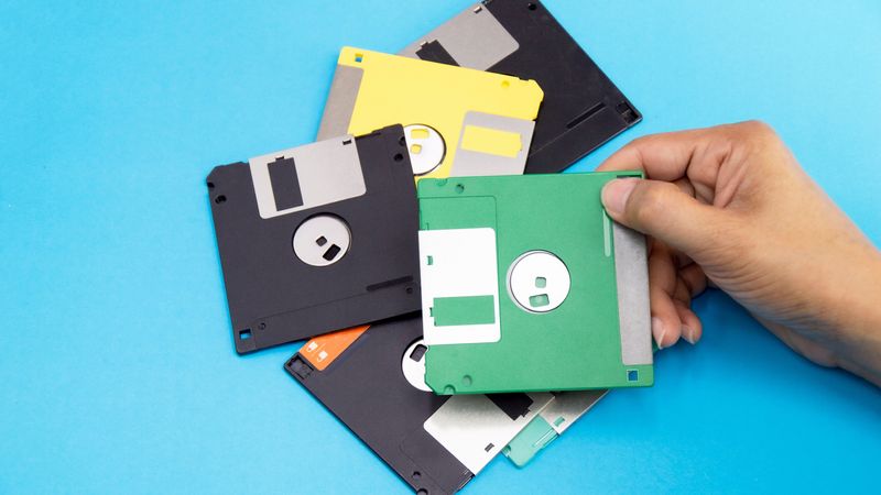 A hand holding one floppy disk with five more on th etable underneath