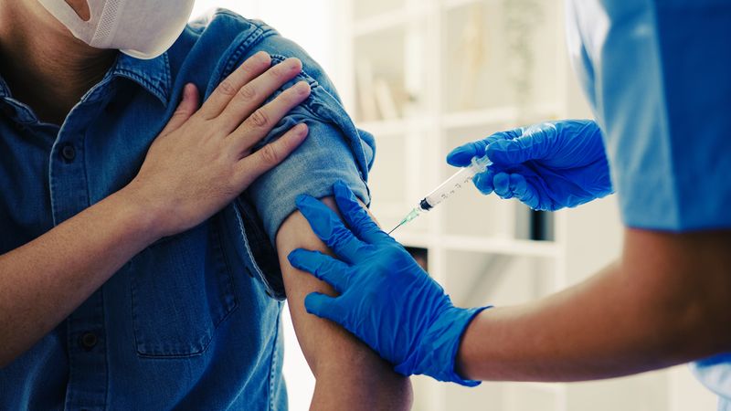 close up of medical professional wearing scrubs and blue gloves administering vaccine into arm of patient wearing blue chambray shirt and white face mask