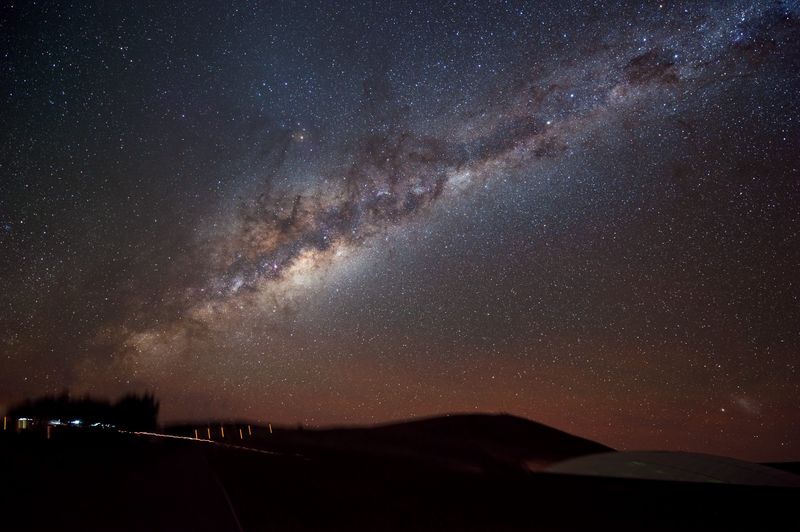 This photograph shows an edge-on view of the Milky Way’s glowing plane slicing across the night sky, laced by bands of dust and dark gas. Taken with a digital camera using a three-minute exposure, the photograph also reveals a bit of action on the ground. To the left, a vehicle with its parking lights on stops lets out a passenger. Though bathed by the light of the Milky Way, the high-altitude desert remains quite dark. To illuminate the rightward path to the underground entrance ramp of the ‘Residencia’, where staff and visitors stay, the passenger takes along a small flashlight, seen as a squiggly bright line. In the lower right, the glass dome on the Residencia’s roof reflects the starry sky overhead. One of our Milky Way’s galactic satellites, the Large Magellanic Cloud, is seen hanging above the Residencia in the lower right corner of the image.