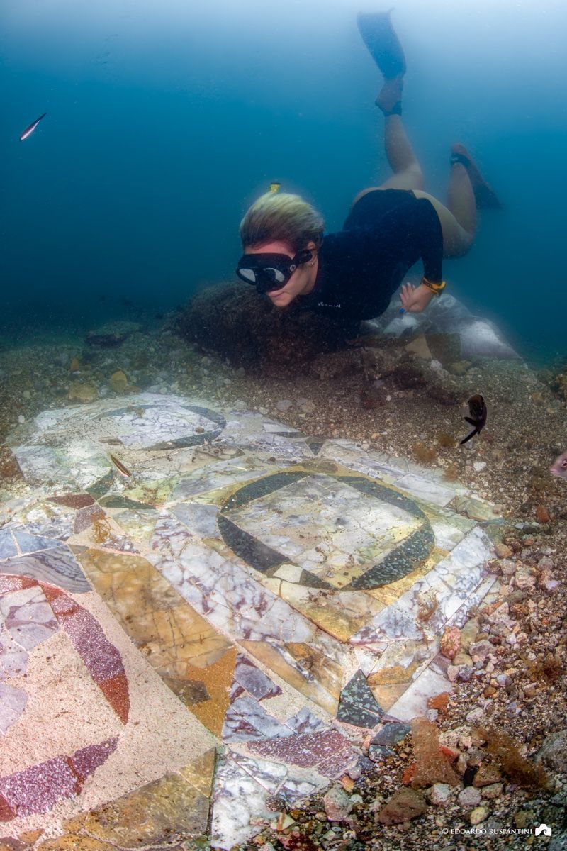 A female diver is looking at part of the marble floor. The floor consists of multiple square marble shapes encircles with rounder stones. There are two of the same blue coloured surrounding and one with a red colour to the left of the image. 