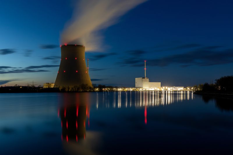 A photo of a cooling tower by a nuclear power plant. The photo is taken from across a lake that is in front of the tower. It appears to be just after sunset, so the tower is set against a clear darkening early night sky. There is a plum of steam rising from the tower and, to the right of the image, the main structures of the power plant itself are illuminated and reflected in the water.