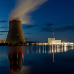 A photo of a cooling tower by a nuclear power plant. The photo is taken from across a lake that is in front of the tower. It appears to be just after sunset, so the tower is set against a clear darkening early night sky. There is a plum of steam rising from the tower and, to the right of the image, the main structures of the power plant itself are illuminated and reflected in the water.