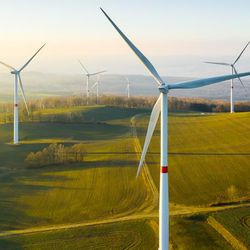 A series of wind turbine are laid out within a network of agricultural fields. The photo is closely focused on one but four others are seen fading into the distance. The sky is clear and it looks like the Sun is still rising. 