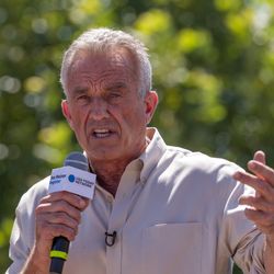 Robert F Kennedy Jr speaking in Des Moines Iowa in August 2023; he is pictured wearing a light beige shirt holding a mic with green trees in the background