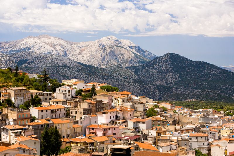 A photo of a town in Sardinia, Italy nestled on a hillside. The red clay tile rooves can be seen sloping from the left to the right of the image. The buildings are all bathed in sunlight. In the background snow covered mountains are visible under a partially clouded sky.