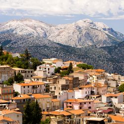 A photo of a town in Sardinia, Italy nestled on a hillside. The red clay tile rooves can be seen sloping from the left to the right of the image. The buildings are all bathed in sunlight. In the background snow covered mountains are visible under a partially clouded sky.