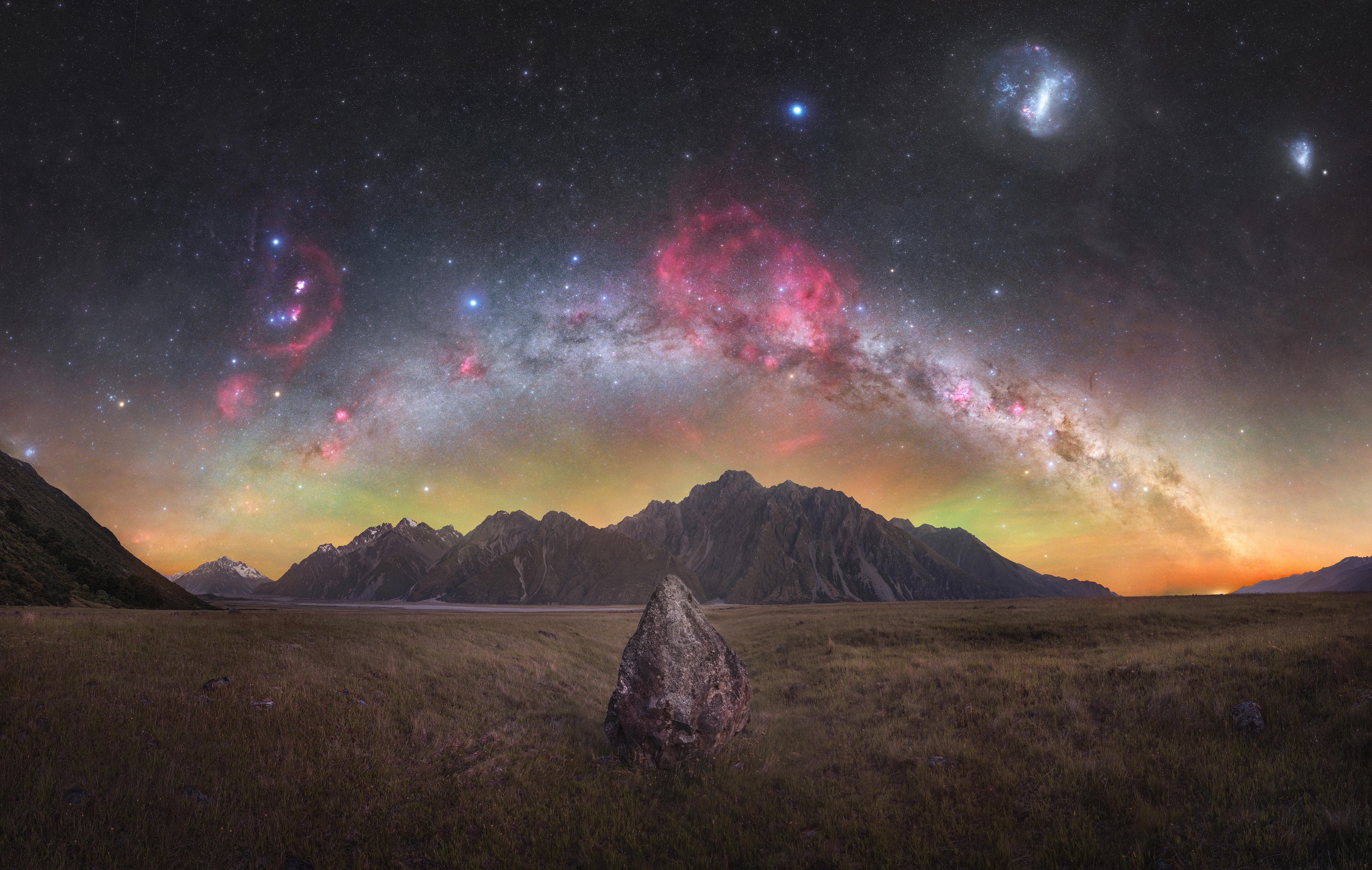 A rock sits on the grounf at the center. mountains are in the background. The milky way arcs above them. The orion constellation and the magelannic clouds are visible above