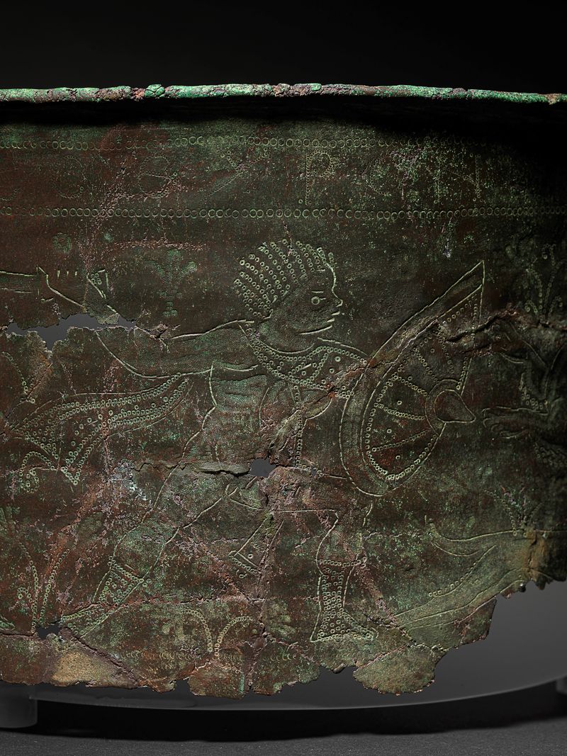 A close up photo of the bronze bucket showing a warrior figure holding a shield and a sword in an outstretched arm. The figure is represented in a two dimensional style. The surface of the bucket is tarnished and dented. 