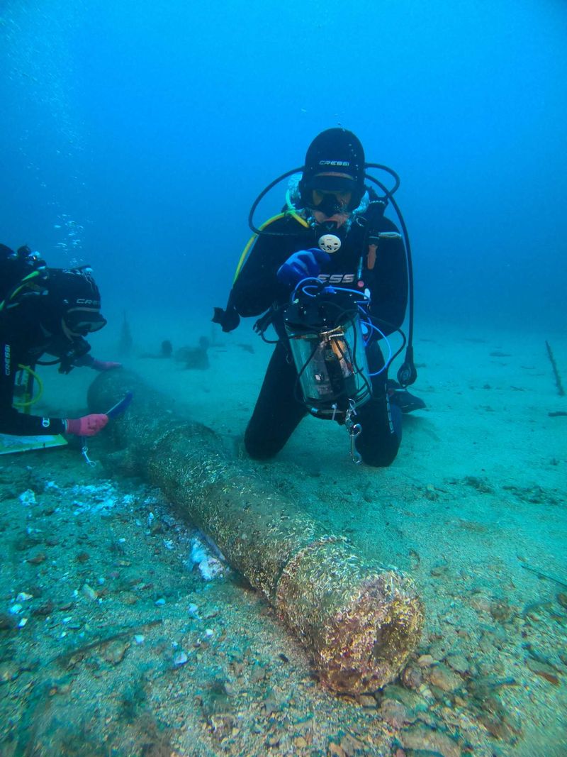 A diver rests on the sea floor in front of the remains of a 16th century cannon. The diver is holding a tubular instrument. Another diver can be seen cleaning end of the cannon in the background. The water visibility is murky and the scene has been lit by an artificial light. 