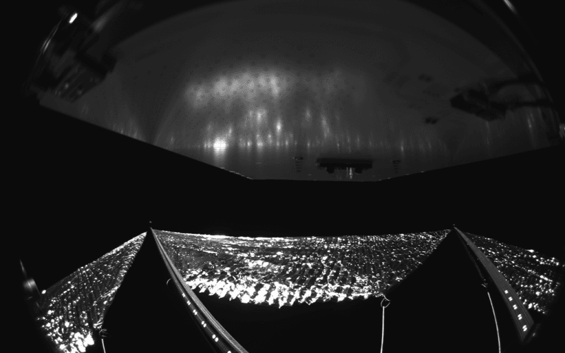  The Advanced Composite Solar Sail System has four black-and-white wide-angle cameras, centrally located aboard the spacecraft. Near the bottom of the photo, the view from one camera shows the reflective sail quadrants supported by composite booms. At the top of the photo is the back surface of one of the spacecraft’s solar panels. The five sets of markings on the booms close to the spacecraft are reference markers to indicate full extension of the sail. The booms are mounted at right angles, and the solar panel is rectangular, but appear distorted because of the wide-angle camera field of view.