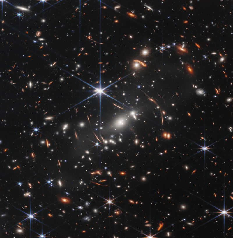 This image shows many overlapping objects at various distances. They include foreground stars, galaxies in a galaxy cluster, and distorted background galaxies behind the galaxy cluster.  The background of space is black. Thousands of small galaxies appear across the image. Their colors vary. Some are shades of orange, others are white. Most appear as fuzzy ovals, but a few have distinctive spiral arms.  In front of the galaxies are several foreground stars. Most appear blue with diffraction spikes, forming eight-pointed star shapes. Some look as large as the galaxies that appear next to them.  A very bright star is slightly off center. It has eight blue, long diffraction spikes. In the center of the image, between 4 o’clock and 6 o’clock in the bright star’s spikes, are several bright, white galaxies.  These are members of the galaxy cluster.  There are also many thin, long, orange arcs. They follow invisible concentric circles that curve around the center of the image. These are images of background galaxies that have been stretched and distorted by the foreground galaxy cluster.