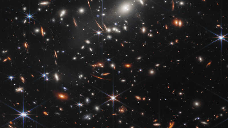 This image shows many overlapping objects at various distances. They include foreground stars, galaxies in a galaxy cluster, and distorted background galaxies behind the galaxy cluster.  The background of space is black. Thousands of small galaxies appear across the image. Their colors vary. Some are shades of orange, others are white. Most appear as fuzzy ovals, but a few have distinctive spiral arms.