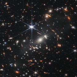 This image shows many overlapping objects at various distances. They include foreground stars, galaxies in a galaxy cluster, and distorted background galaxies behind the galaxy cluster.  The background of space is black. Thousands of small galaxies appear across the image. Their colors vary. Some are shades of orange, others are white. Most appear as fuzzy ovals, but a few have distinctive spiral arms.  In front of the galaxies are several foreground stars. Most appear blue with diffraction spikes, forming eight-pointed star shapes. Some look as large as the galaxies that appear next to them.  A very bright star is slightly off center. It has eight blue, long diffraction spikes. In the center of the image, between 4 o’clock and 6 o’clock in the bright star’s spikes, are several bright, white galaxies.  These are members of the galaxy cluster.  There are also many thin, long, orange arcs. They follow invisible concentric circles that curve around the center of the image. These are images of background galaxies that have been stretched and distorted by the foreground galaxy cluster.