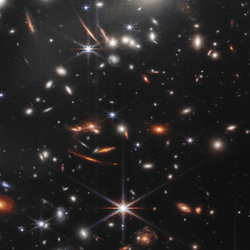 This image shows many overlapping objects at various distances. They include foreground stars, galaxies in a galaxy cluster, and distorted background galaxies behind the galaxy cluster.  The background of space is black. Thousands of small galaxies appear across the image. Their colors vary. Some are shades of orange, others are white. Most appear as fuzzy ovals, but a few have distinctive spiral arms.