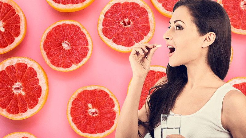 woman with long dark hair taking a pill holding a glass of water, against a backgroun of a repeating pattern of pink grapefruit slices against a light pink background