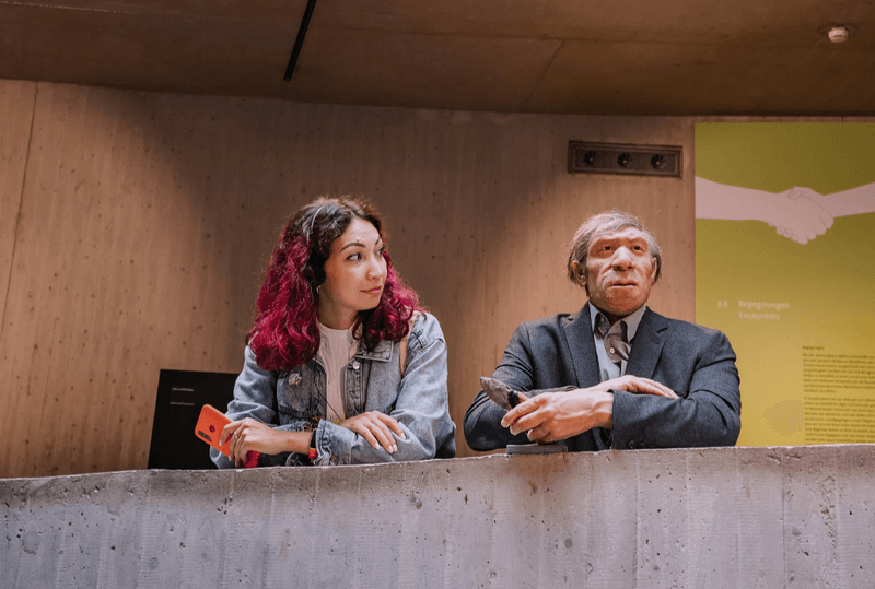 Homo sapiens (human) standing next to a model of a Neanderthal dressed like a modern human. 