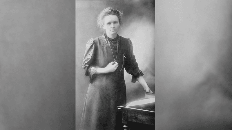 black and white photo of Marie Curie standing by a desk holding a pendant