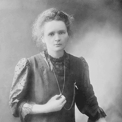 black and white photo of Marie Curie standing by a desk holding a pendant