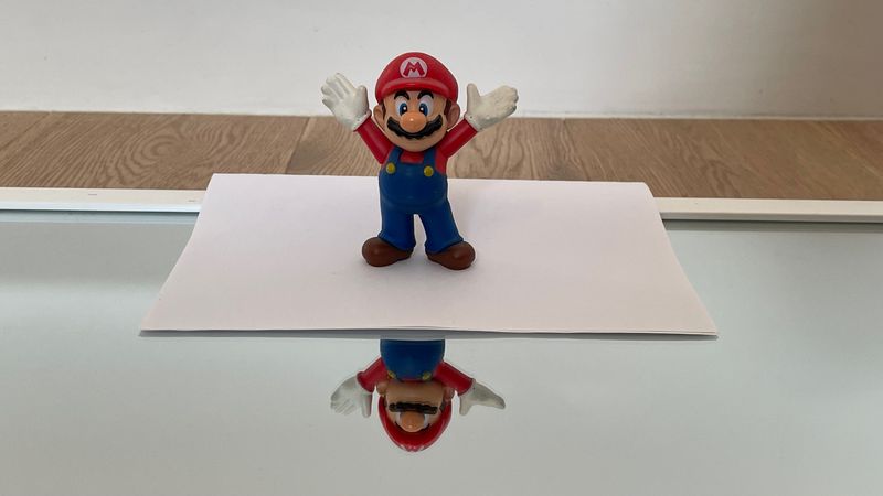 A Mario statuette sits on a piece of paper on a mirror, which should be hidden from the mirror by the paper. Given the angle, however, part of the statue is visible in the mirror.