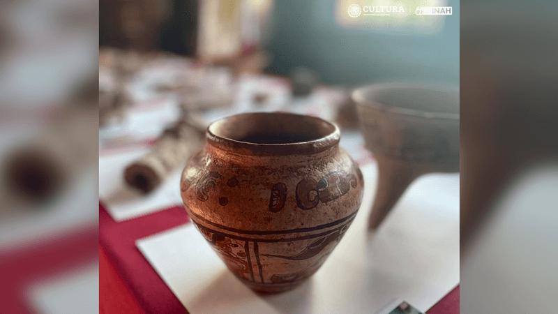 The Maya vase sits on a square of white paper on a table covered with a burgundy cloth. The vase has a tea-stained colour to it and its body has images painted onto its surface in rust red colour. The images are difficult to make out in the photo, but they represent humanoid figures and some design patterns.