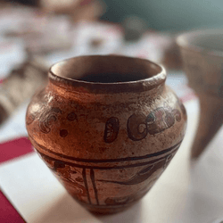 The Maya vase sits on a square of white paper on a table covered with a burgundy cloth. The vase has a tea-stained colour to it and its body has images painted onto its surface in rust red colour. The images are difficult to make out in the photo, but they represent humanoid figures and some design patterns.