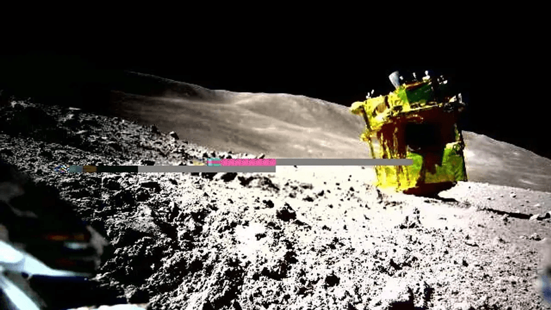 SLIM as it is currently on the surface of the Moon, seen by one of its rovers.  The golden lander is on its side with the rockets pointing up and the solar panels in shade