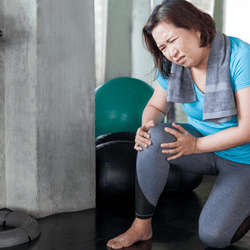 Woman kneeling in a gym clutching her knee in pain, wearing a blue T shirt and grey leggings with a towel around her shoulders